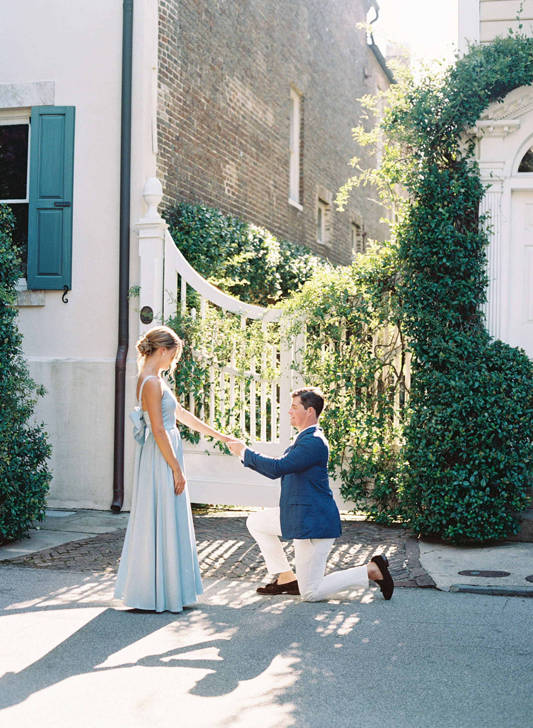 Man proposing to a woman in a light blue dress outdoors.