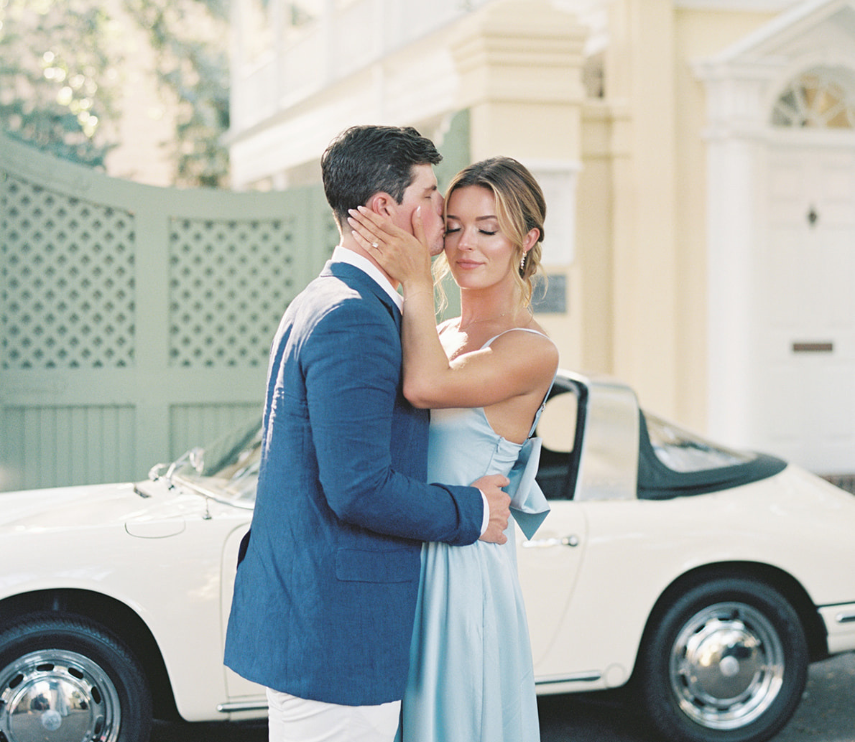 Couple embracing in front of a vintage car with a building in the background